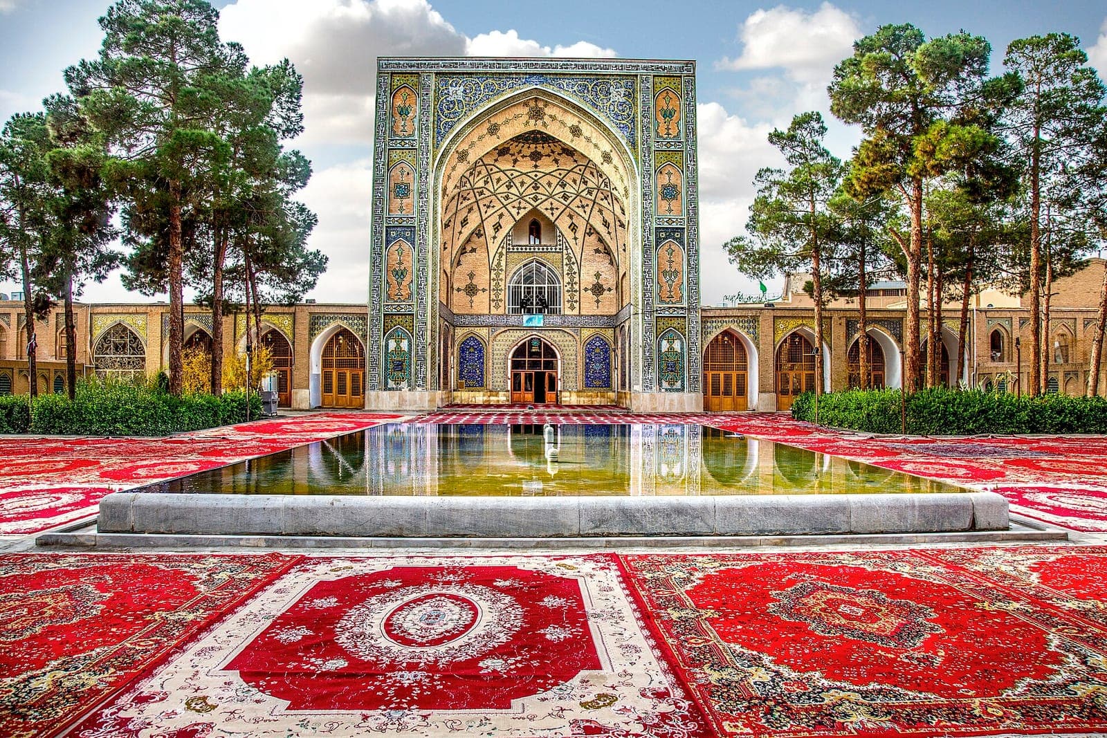 ایوان کاشیکاریشدهٔ مسجد امام (سلطانی) سمنان با حوض میانی، درختان کاج و فرشهای سرخ گسترده در صحن
