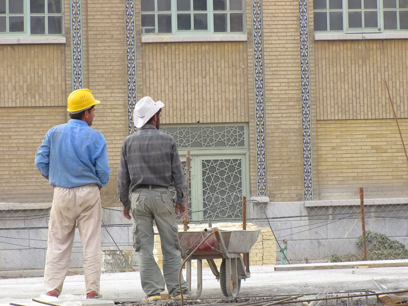 دو کارگر ساختمانی با کلاه ایمنی، از پشت سر، در کارگاه ساخت یک مسجد با نمای آجری و کاشیکاری